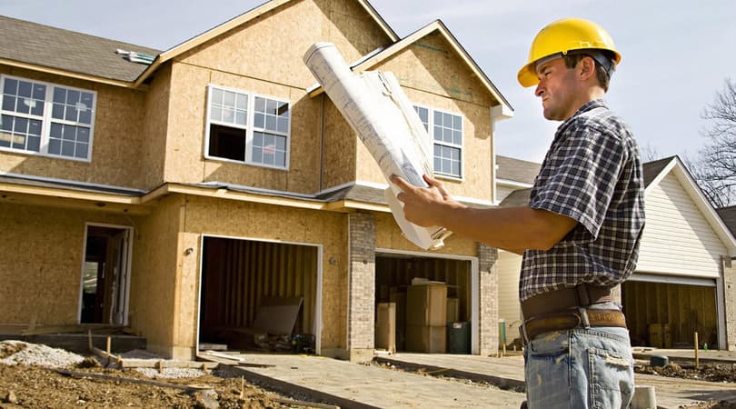 A construction manager in a hard hat reviewing blueprints outside a new home