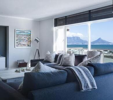 A bright, modern living room with a large blue sectional sofa, a coffee table, and a wide window overlooking the ocean and mountains.