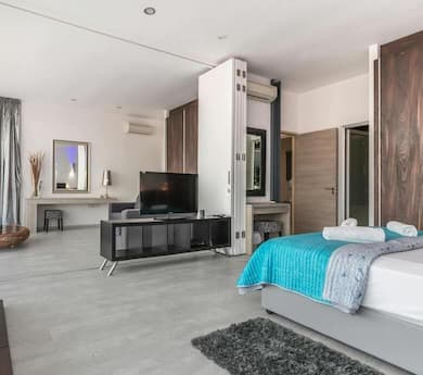 A spacious and modern bedroom with a bed in the foreground, a TV and entertainment unit, a dressing table with a mirror, and an open doorway leading to another area.